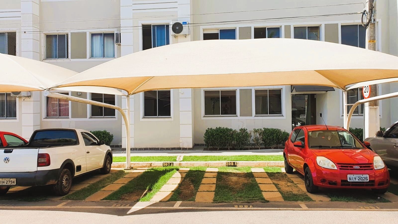 Un área de estacionamiento con dos coches bajo una marquesina, un edificio al fondo y una señal de límite de velocidad.