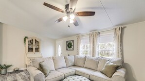 Una sala de estar con un sofá seccional, un otoman redondo, un ventilador de techo, un mueble aparador y una ventana con cortinas.