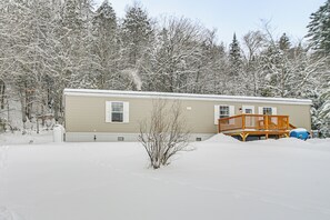 A single-story mobile home with a wooden deck, surrounded by snow-covered trees.