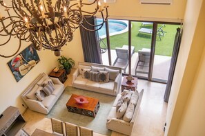 A spacious living area with a large sofa set, a coffee table, and a chandelier. There is a view of a pool and outdoor seating through large glass doors. - Spacious 4-bedroom villa in fantastic Cabo San Lucas with AC (Cabo San Lucas)