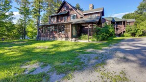 Une cabane rustique en rondins de bois, dotée d’un grand porche à l’avant, entourée d’une végétation luxuriante et de grands arbres.