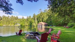 Un chalet au bord du lac, doté d’un foyer, de chaises Adirondack rouges et offrant une vue sur le lac et la forêt.
