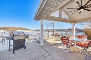 Eine überdachte Terrasse mit Grill, Sitzgelegenheiten im Freien und Blick auf See und Hügel.