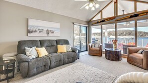A spacious living area with a gray sofa, a brown leather chair, and a round coffee table. There is a large window providing a view of the outdoors. - Private Hot Tub, Fire Pit! Lakefront Ozarks Escape (Gravois Mills)