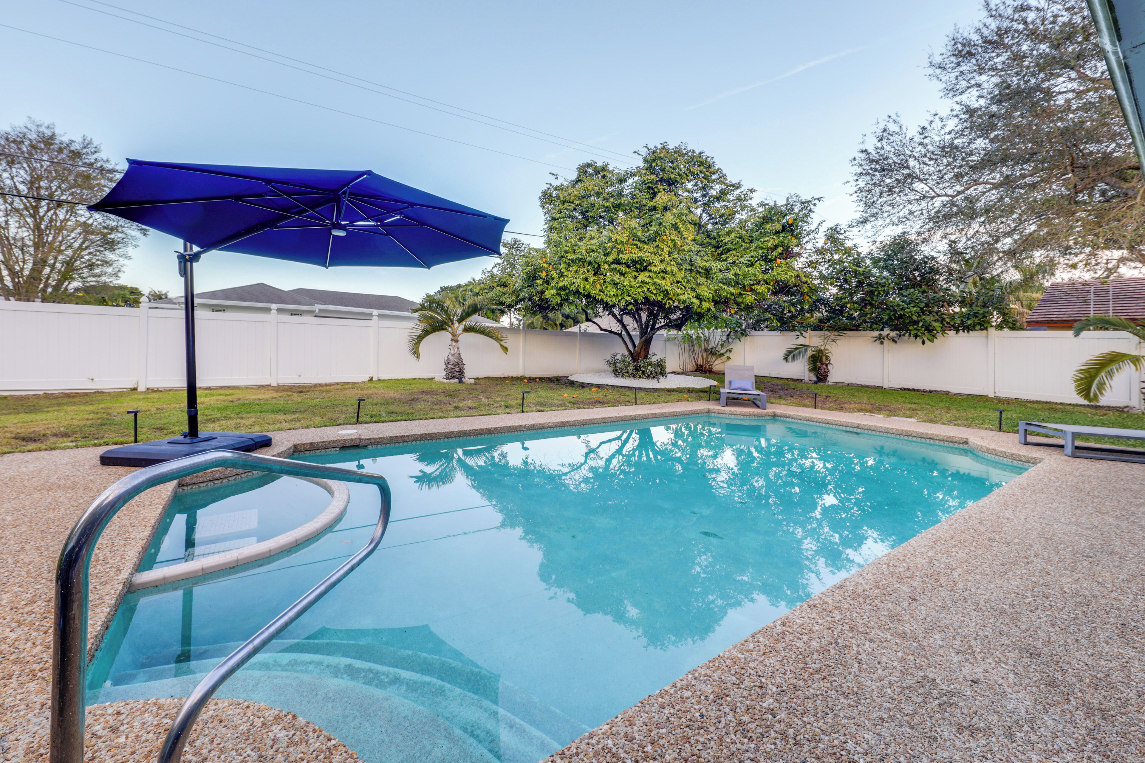 A swimming pool with a blue umbrella, surrounded by a white fence, palm trees, and a clear sky.