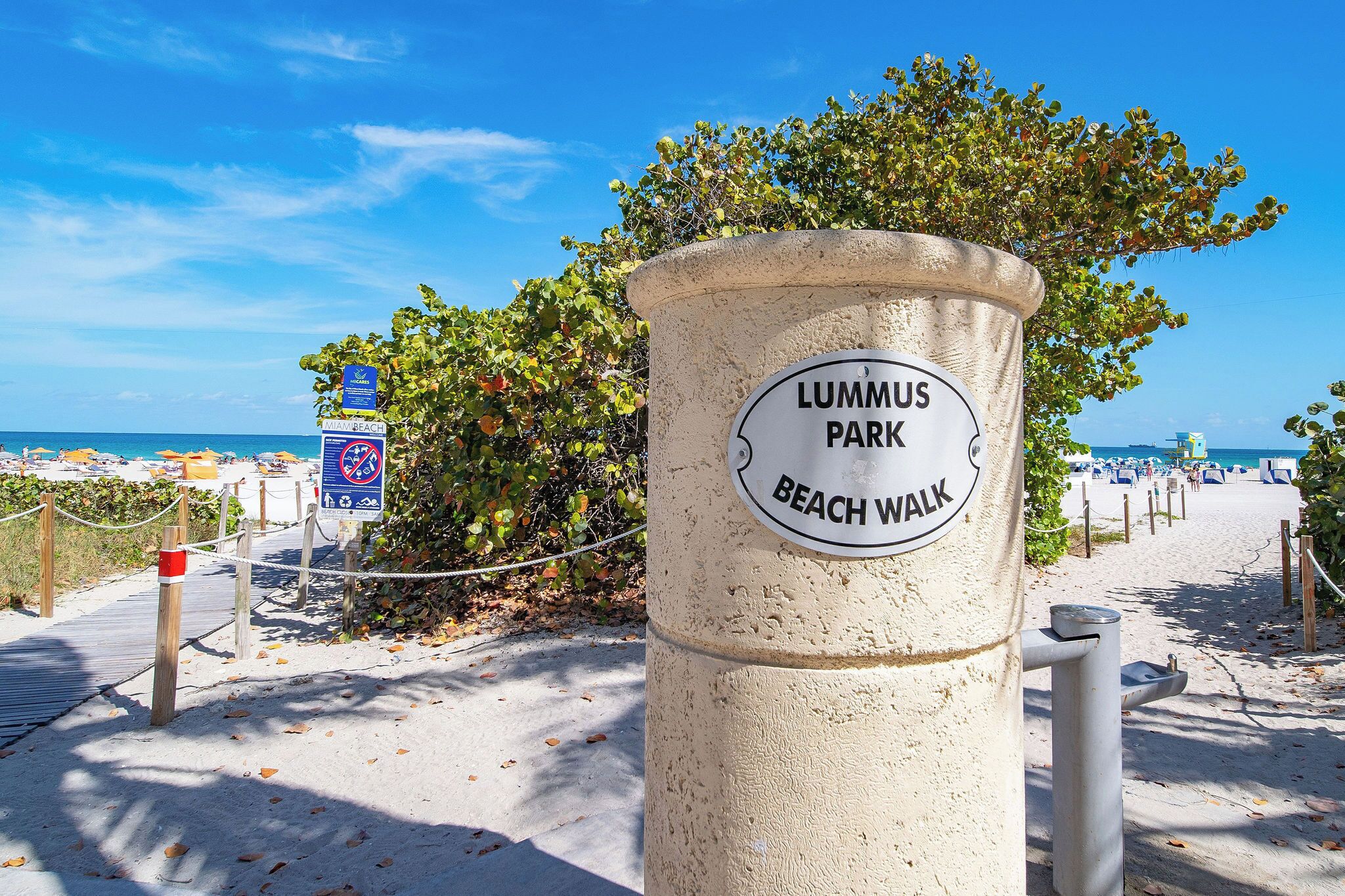 A sign for Lummus Park Beach Walk with a clear view of the beach and ocean in the background.