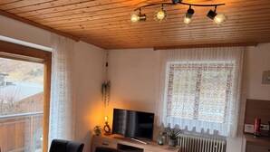 Una habitación acogedora con techo de madera, un televisor sobre un mueble de madera, un radiador y una ventana con cortinas de encaje.