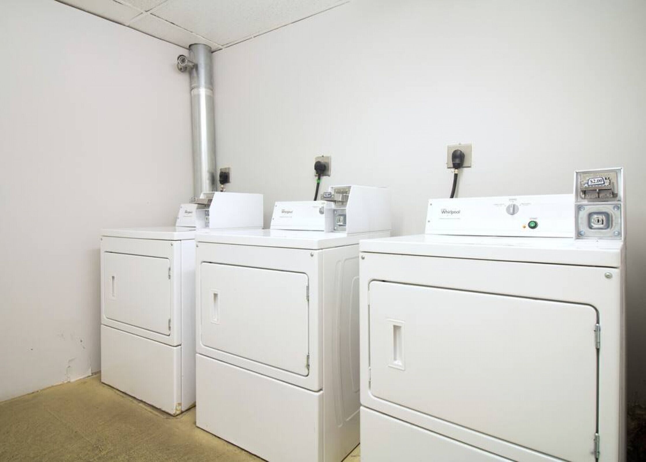 A laundry room with two white Whirlpool washers and dryers, a metal vent, and a power outlet.