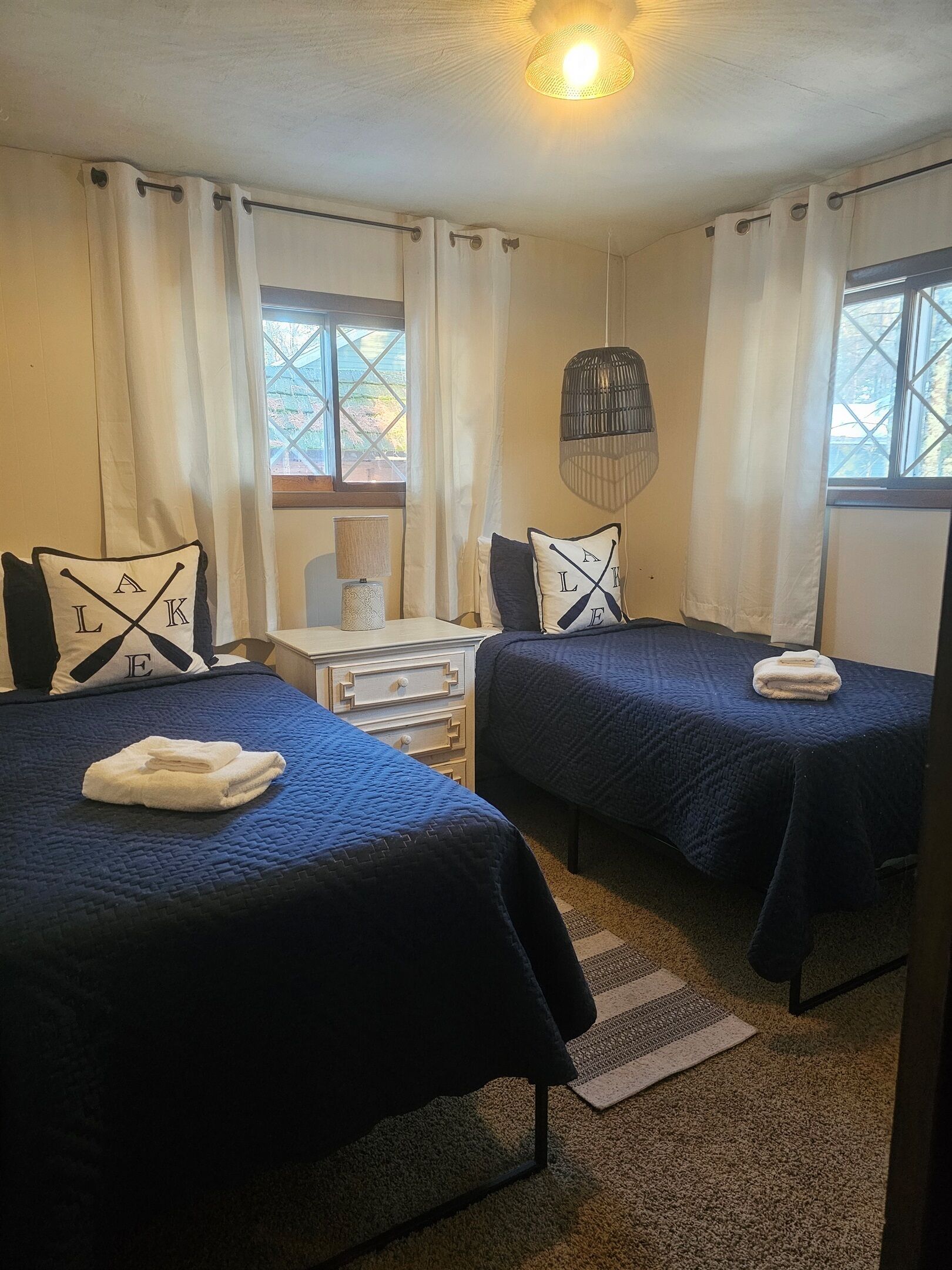 A hotel room with two beds, a small table, a lamp, and a view of the outdoors through the windows.
