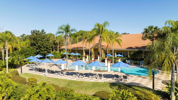 Ein Poolbereich eines Resorts mit Liegestühlen und Sonnenschirmen, umgeben von Palmen und einem Gebäude mit einem Ziegeldach.