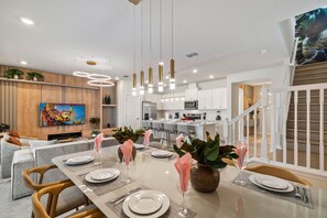 A modern dining area with a long table set for six, a kitchen in the background, and a staircase leading to an upper level. - Designer 5BR GameLoft, Foam Pit & Lazy River 16164 (Clermont)