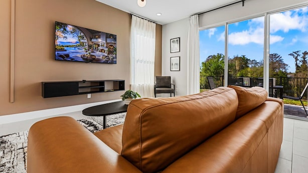 Ein modernes Wohnzimmer mit einer Ledersofa, einem Flachbildfernseher an der Wand, einem kleinen Tisch mit Pflanze und einem Balkon mit Gartenmöbeln.