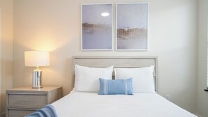 Un lit bien fait, avec du linge de lit blanc et des touches de bleu, une lampe de chevet et deux tableaux encadrés accrochés au mur.
