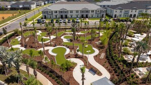 Vue aérienne d’un quartier résidentiel avec un jardin paysager central, agrémenté de chemins sinueux et de palmiers, le tout entouré d’immeubles d’appartements modernes.
