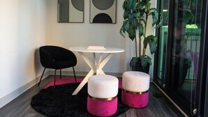 Una habitación moderna con una mesa redonda blanca, dos otomanas rosas, una silla negra y dos obras de arte abstracto en la pared.