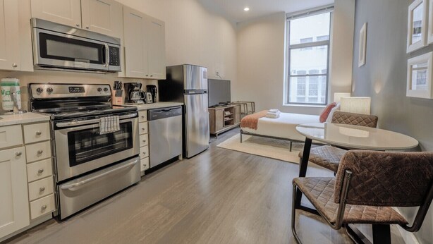 Cocina compacta con electrodomésticos modernos, zona de comedor con mesa y sillas, y cama con vistas a la ciudad.