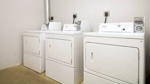 Zona de lavandería con tres lavadoras y secadoras Whirlpool blancas, un conducto de ventilación metálico y enchufes en la pared.