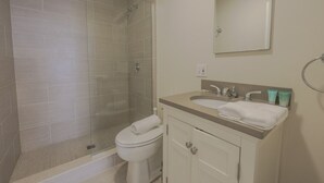 Un baño con ducha envuelta en vidrio, inodoro blanco, lavamanos con dos grifos y espejo sobre el lavamanos.