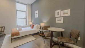 Una sala de estar moderna con sofá cama, una mesa redonda y dos sillas. Hay una ventana que proporciona luz natural y una pared con cuadros.