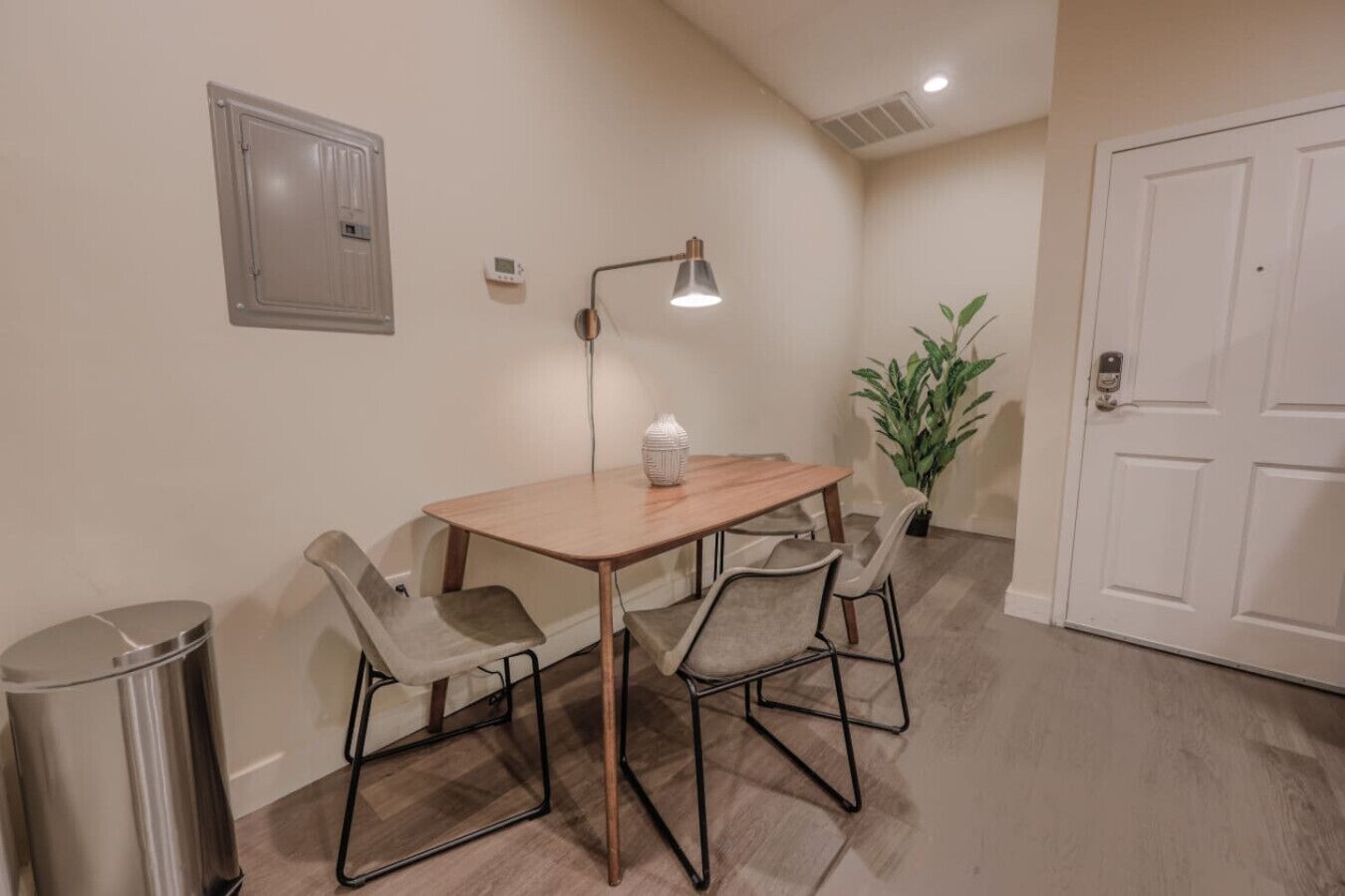 A small dining area with a table and four chairs, a trash can, a wall-mounted light, a potted plant, and a door.