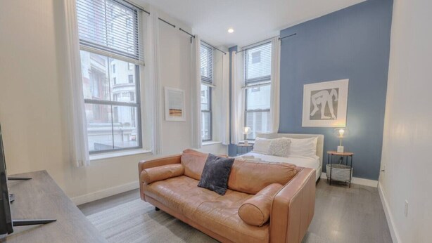 A modern bedroom with a large window, a bed, a sofa, and a bedside table with a lamp. - Pet Friendly Studio | Walk to MGH (Boston)