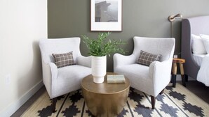 Quarto de hotel moderno com duas poltronas cinzas, uma mesa de centro redonda de madeira com uma planta, um quadro na parede e um tapete estampado.