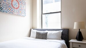 A neatly made bed with white bedding, a gray headboard, and a patterned pillow. A bedside table with a lamp is visible to the right. - Center of Boston Studio Pet Friendly | Laundry (Boston)
