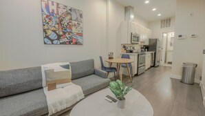 Een modern appartement met een grijze bank, een ronde salontafel met een plant, een keuken met witte kastjes en een kleurrijk stripboek aan de muur.