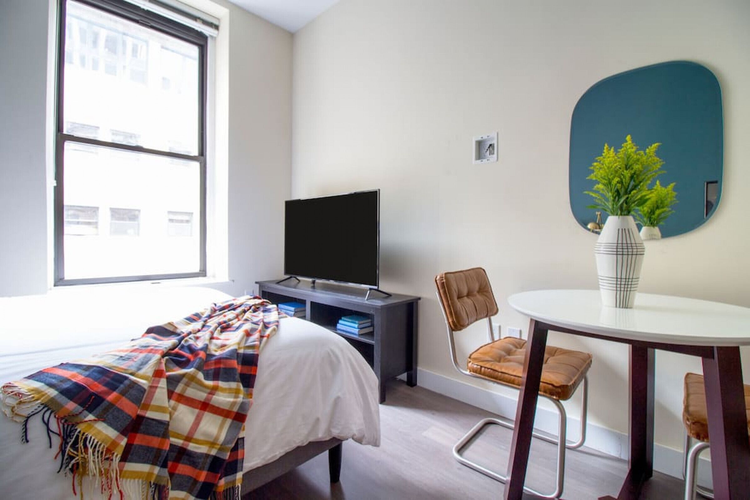 Habitación de hotel con cama, televisor sobre un soporte, una mesa de comedor con dos sillas y un ventanal que ofrece vistas a edificios.