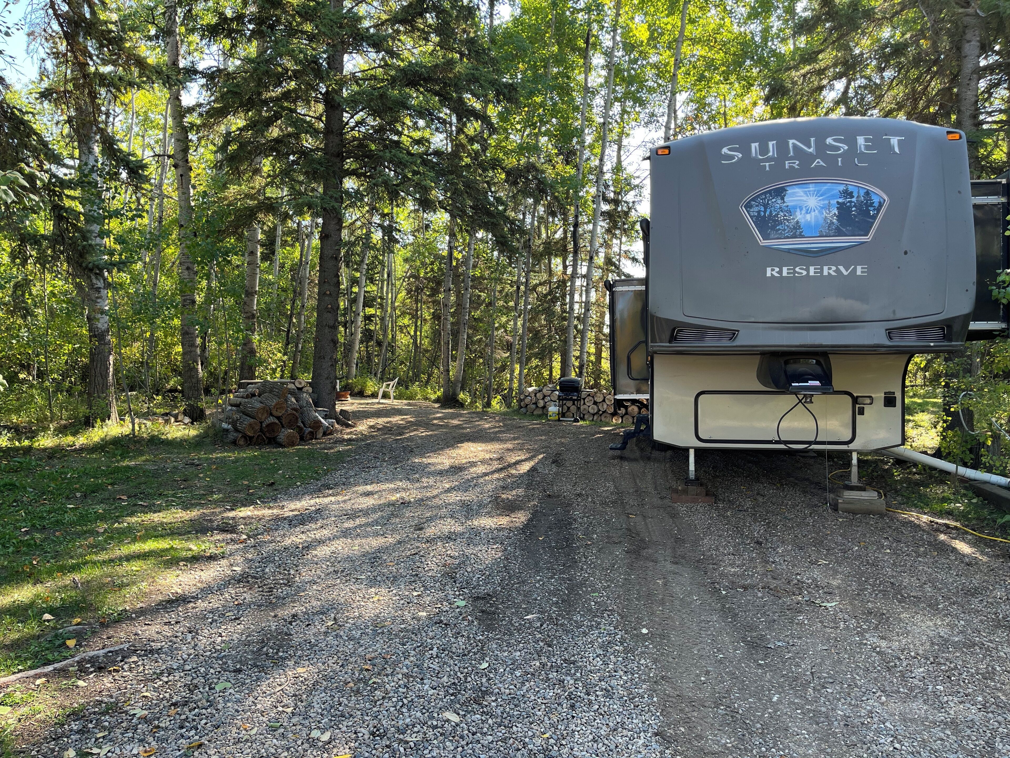 Un terrain de camping où est stationné un grand camping-car, baptisé « SUNSET TRAIL RESERVE », sur un chemin de gravier, entouré d’arbres.