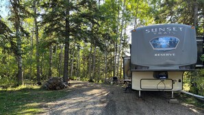 A campsite with a large RV named 'SUNSET TRAIL RESERVE' parked on a gravel path surrounded by trees.