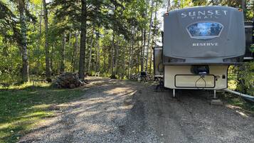 Un terrain de camping où est stationné un grand camping-car, baptisé « SUNSET TRAIL RESERVE », sur un chemin de gravier, entouré d’arbres.