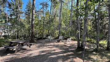 Un terrain de camping situé dans une forêt, équipé d’une table de pique-nique et de deux chaises en plastique blanc.