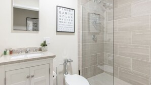 Un baño con un lavamanos blanco, un espejo y un inodoro. Cuenta con una ducha con puertas de vidrio y un cartel enmarcado en la pared.