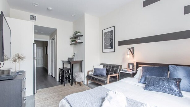 Habitación de hotel moderna con una cama grande, un escritorio, una silla y una mesita pequeña. Se ve una puerta que da a otra habitación y una repisa en la pared con objetos decorativos.