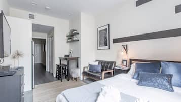 Ein modernes Hotelzimmer mit einem großen Bett, einem Schreibtisch, einem Stuhl und einem kleinen Tisch. Eine Tür führt in einen weiteren Raum, und an der Wand befindet sich ein Regal mit Dekoration.