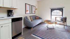 A modern kitchen with white cabinets, a coffee maker, and a dishwasher. A gray sofa with patterned cushions is against the wall, and a wooden coffee table is in the foreground. - Downtown Boston | Nurses Welcome | W/D in bldg (Boston)