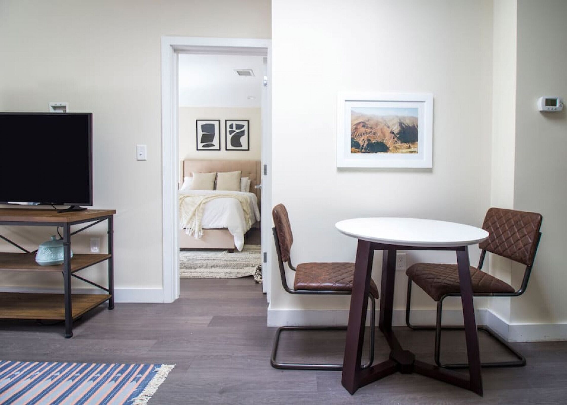 Une chambre d’hôtel moderne dotée d’un téléviseur à écran plat posé sur un meuble en bois, d’un coin repas avec une table ronde et deux chaises, et d’un lit avec une tête de lit beige et une literie blanche.