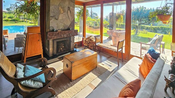 A spacious living room with a stone fireplace, large windows, and a view of a pool and garden. - w*| Spacious 4BR Country House with Pool in Mala (Asia)