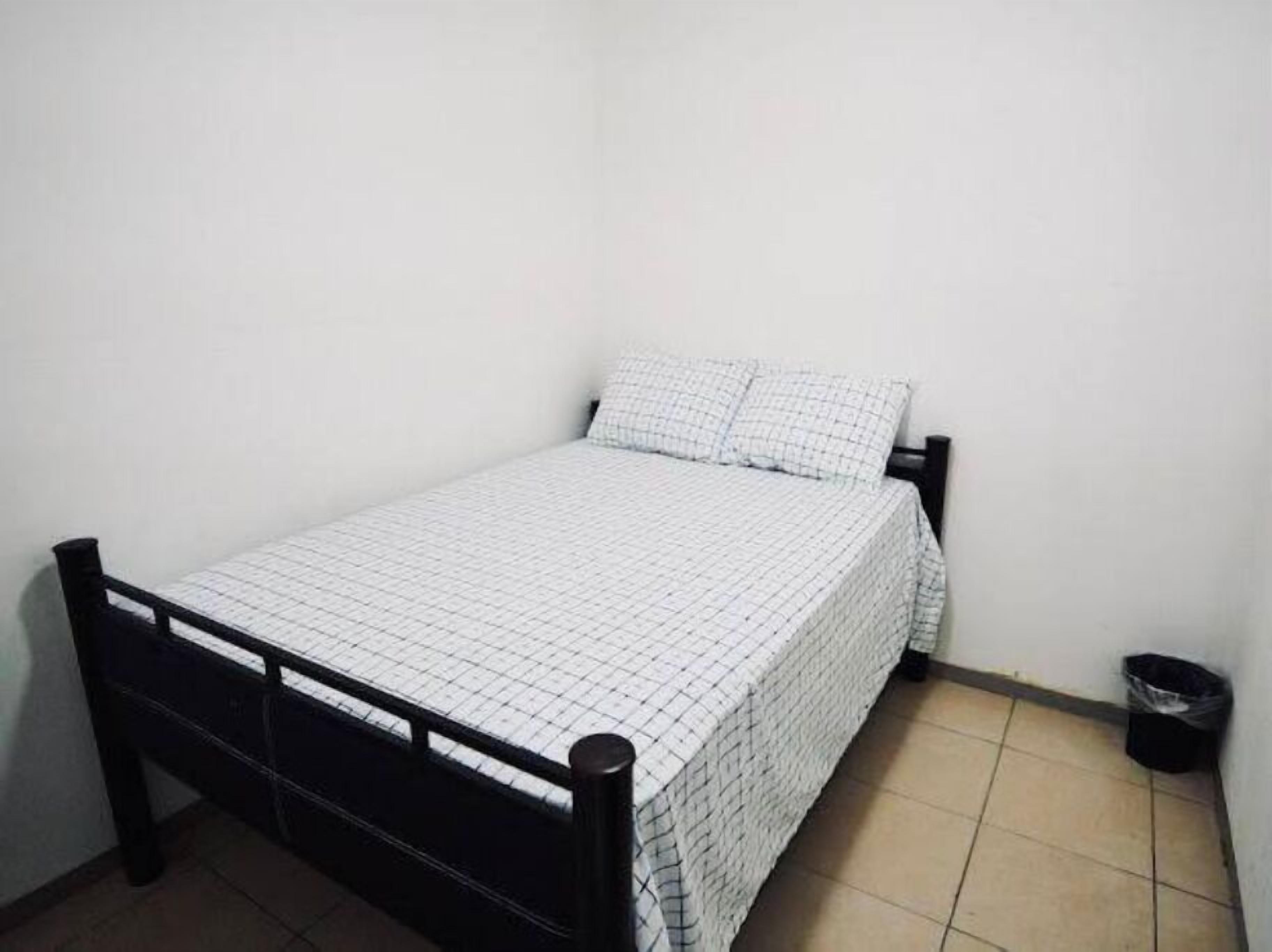 Una habitación pequeña con cama individual, una pared blanca lisa, estructura de metal negro y colcha blanca a cuadros.