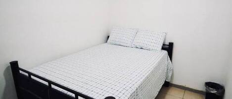 Una habitación pequeña con cama individual, una pared blanca lisa, estructura de metal negro y colcha blanca a cuadros.