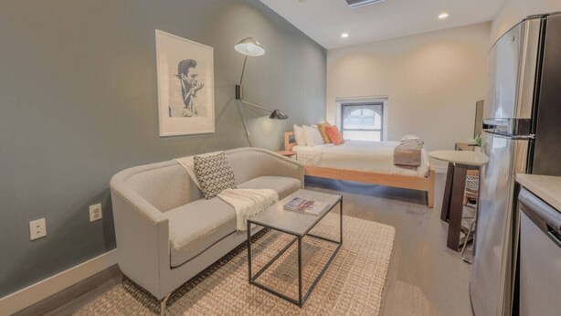 A compact, modern room with a sofa, a small table, a bed, a kitchenette, and a ceiling light. - Serene Studio | Close to MGH (Boston)