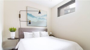 Ein ordentlich bezogenes Bett mit weißen Leinen, ein Kopfteil mit einer gepolsterten Struktur, ein an der Wand hängendes Bild von Bergen, ein kleiner Nachttisch mit einer Pflanze und eine Nachttischlampe.