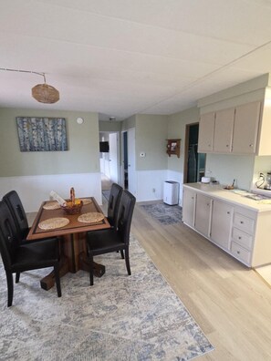 A dining area with a wooden table and chairs, a kitchen with white cabinets, and a hallway leading to another room. - Coastal Retreat With Bay Views (Eureka)