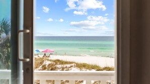 A beach view from a window with a clear sky, a sandy beach, and a few people and umbrellas.