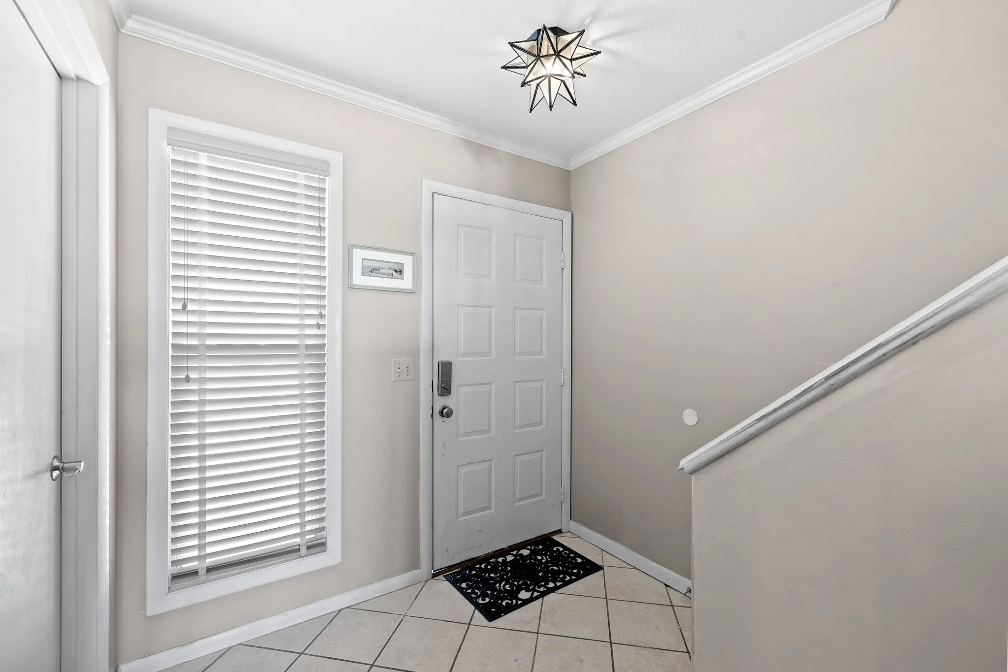 A hallway with a white door, a window with blinds, a black doormat, and a ceiling light fixture.