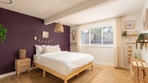 Een slaapkamer met een houten bed, een raam met gordijnen, een nachtkastje en een wandplank met planten.