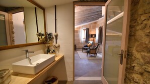Une salle de bain moderne avec un grand miroir, un lavabo blanc et un meuble-lavabo en bois. Une porte donne sur une chambre à coucher équipée d’un lit et d’une chaise.