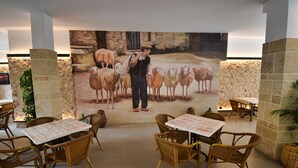 Un restaurant avec des chaises et des tables en osier, une fresque représentant un homme avec des moutons, et des murs en pierre.