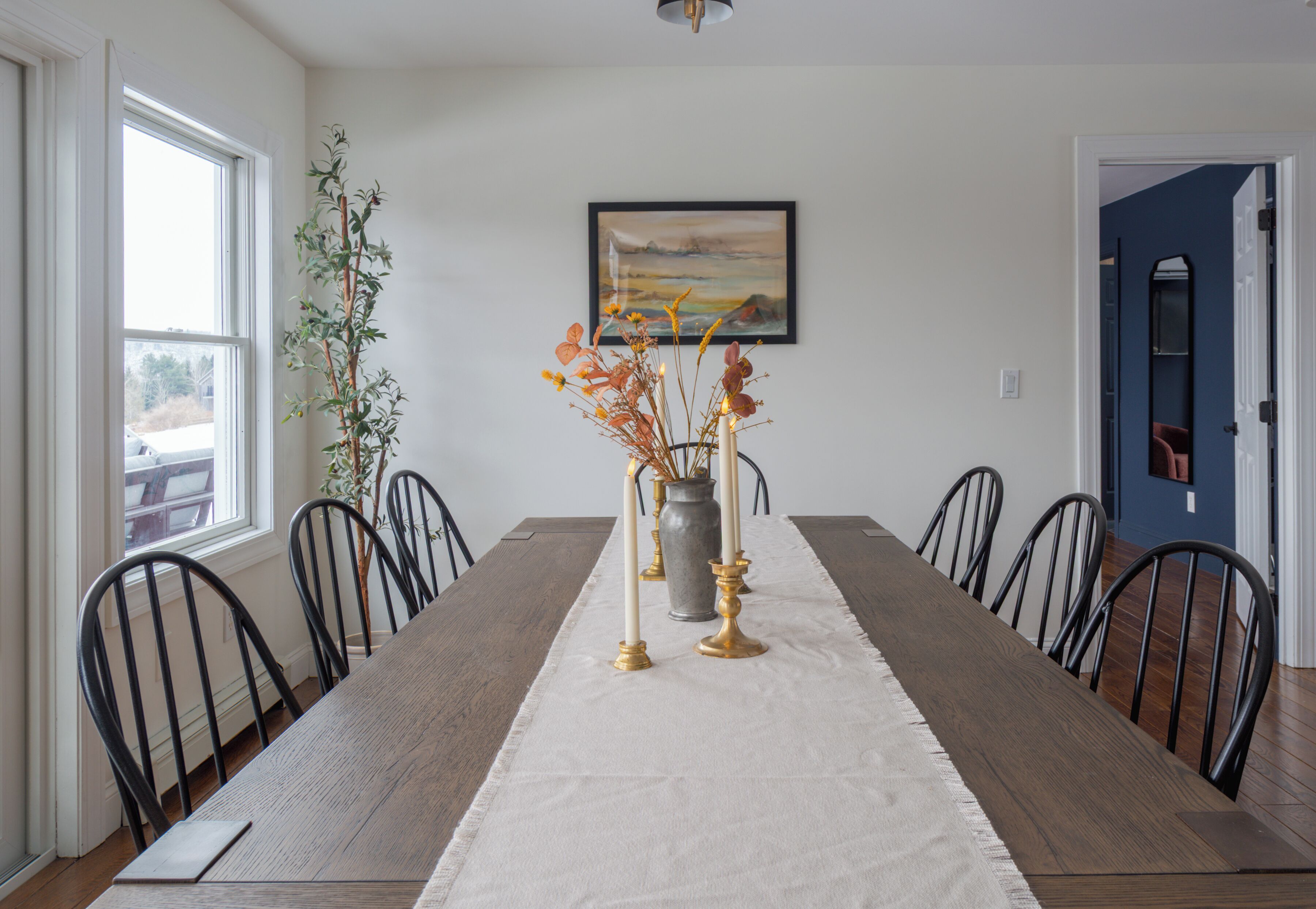 Un comedor con una mesa larga, sillas, un jarrón con flores y un cuadro en la pared.